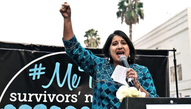 Sonali Kolhatkar raises her right hand in a fist while speaking into a microphone she holds in the other in front of a #MeToo Survivors March banner.