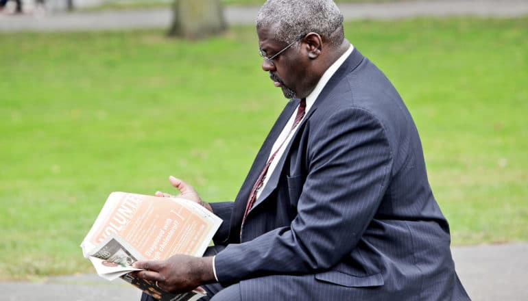 African American man in navy suit reads newspaper on outdoor bench