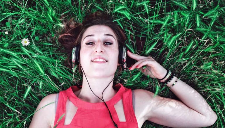 woman lies in grass with one arm raised to hold silver headphones on her head - listening to podcasts