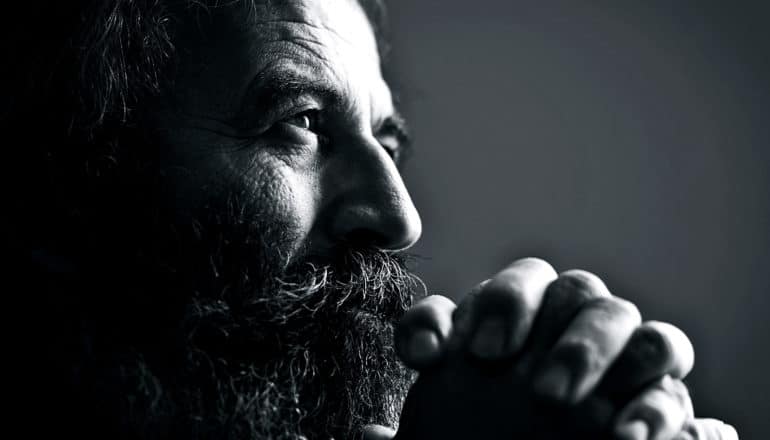 A bearded man clasps his hands together in prayer in black and white.