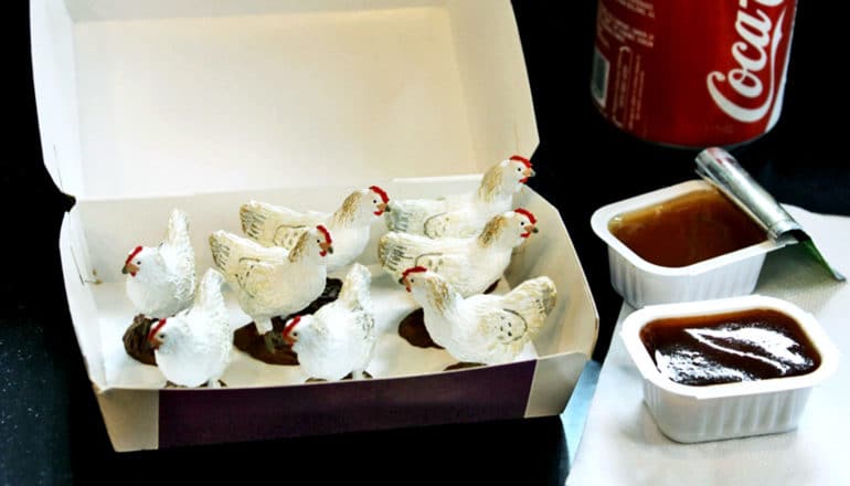 Small toy chickens sit in a food container next to some dipping sauces and a canned soda