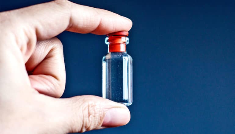 A hand holds a tiny glass bottle with a red lid between the thumb and index finger.