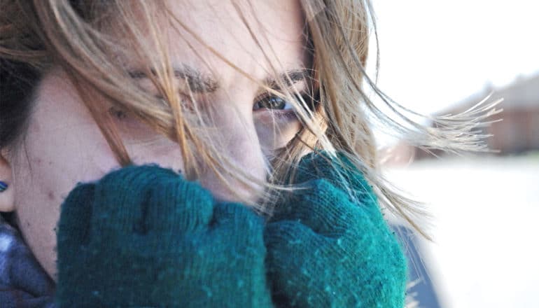 A young woman holds green wool gloves to her face as the wind blows her hair