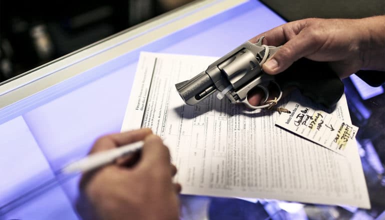 The image shows a man filling out background check paperwork with a gun in hand. (Universal background checks concept)