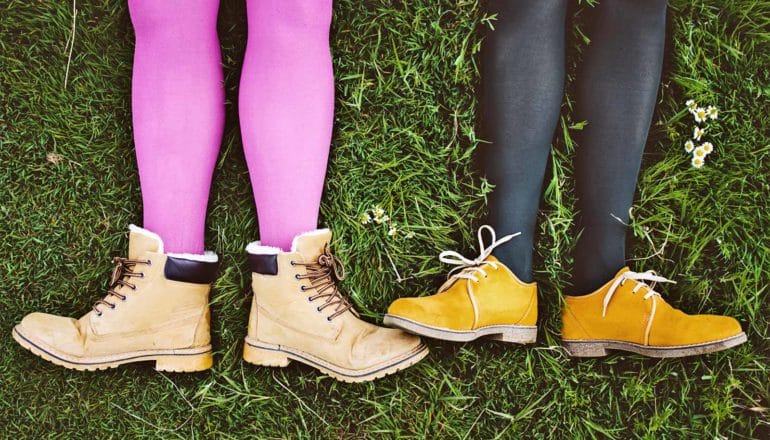 Two pairs of legs in nylon leggings lay in the grass, one with boots and the other with yellow shoes.