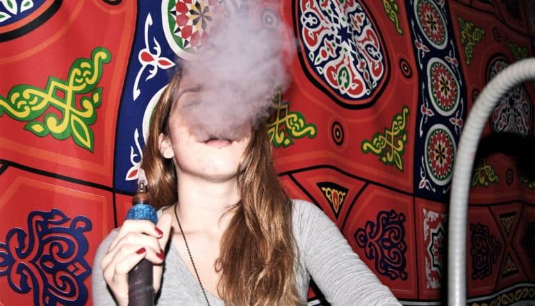 The image shows a young woman smoking hookah with a wall tapestry behind on the wall behind her. (hookah concept)