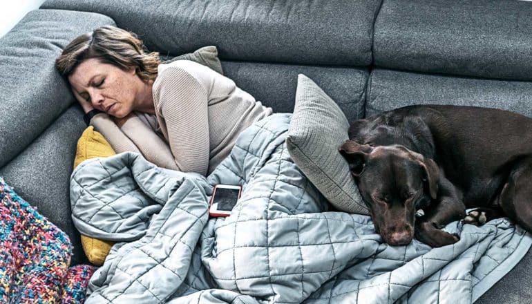 A woman sleeps on the couch under a blanket while her dog naps at her feet.