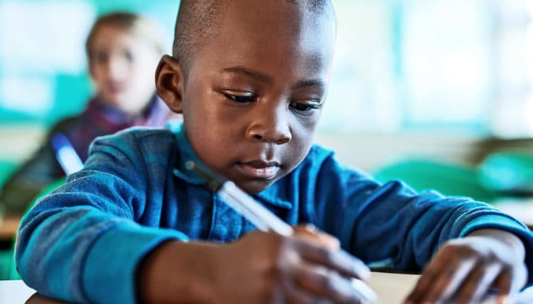 The image shows a young boy in a classroom looking down at his paper while holding a pen. (gifted kids concept)