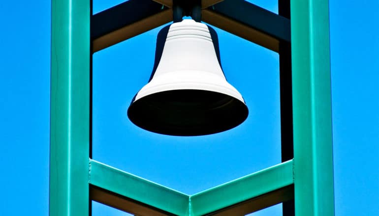 A bell hangs in a green metal structure against a blue sky, with the bell's hole appearing totally black