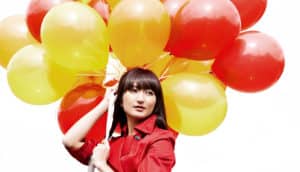 A woman in a red coat holds a huge bunch of yellow, orange, and red balloons