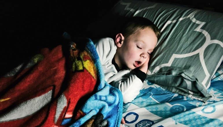 A young boy sleeps soundly in his bed under a colorful blanket with his mouth open