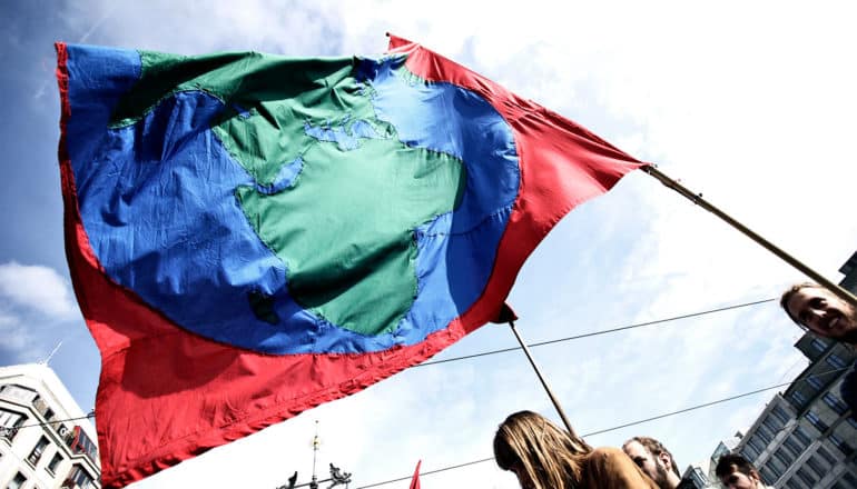 A protestor holds up a red flag with an image of the earth on it, which waves against a cloudy sky