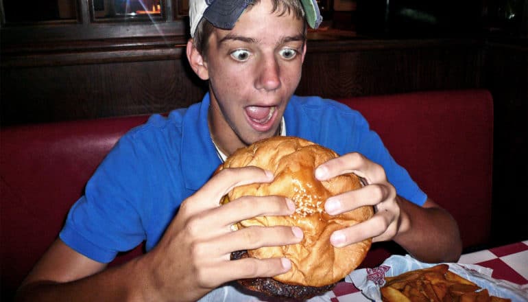 A young man in a blue shirt and ball cap looks in awe at a very large hamburger