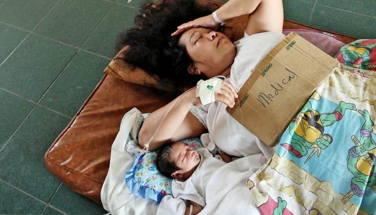 woman and newborn baby lie on cushion. Woman holds cardboard sign saying "medical" and holds hand to head