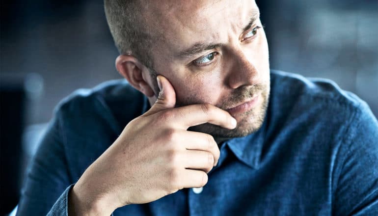 An man in a blue shirt puts his hand to his face while he's deep in thought, looking off to his left