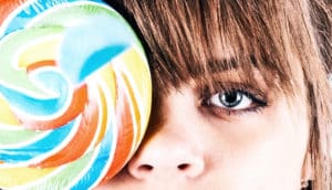 A young woman holds a colorful, rainbow-swirled lollipop over her right eye