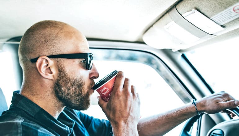 A man wearing sunglasses drives to work while sipping a cup of coffee