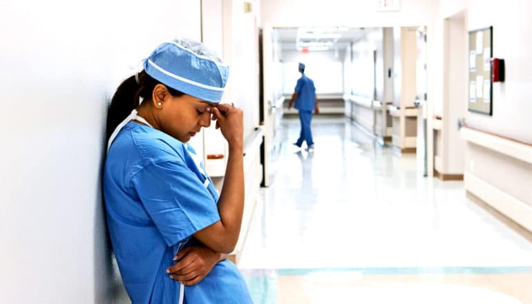 A doctor in scrubs leans against a hospital hallway wall, touching her hand to the bridge of her nose as a male colleague can be seen in the background walking away