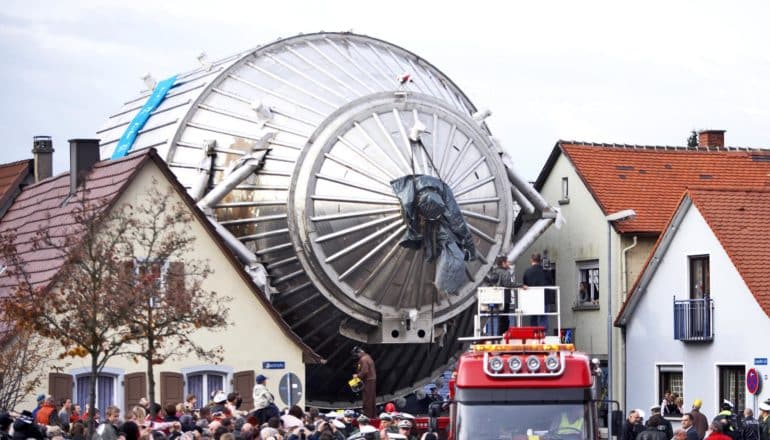 huge piece of scientific equipment passes between houses amid crowd