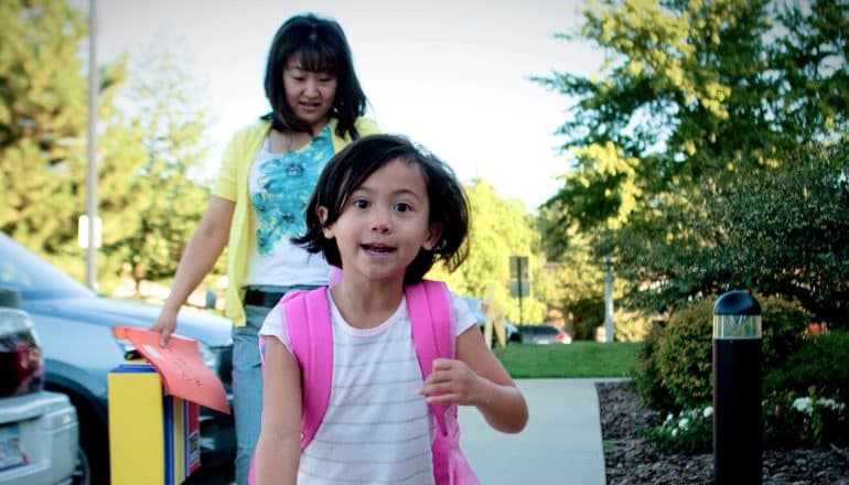 excited child runs toward camera with mother behind carrying folder