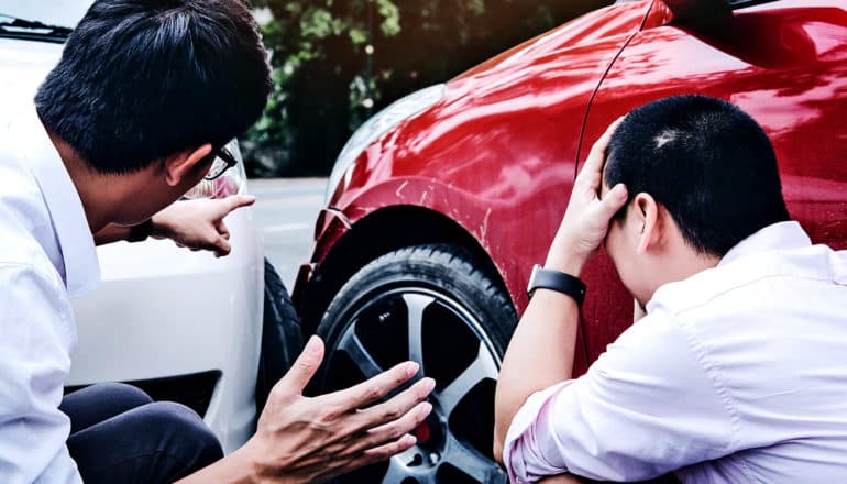 two men kneel by their cars after a crash, with one pointing to the damage and the other holding his head in his hands.