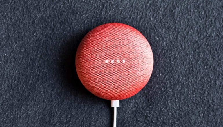 A red, circular Google home speaker sits on a dark surface with a USB power cord coming out of the bottom