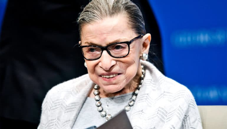 Ruth Bader Ginsburg smiles as she's speaking to a group while against a black and blue background