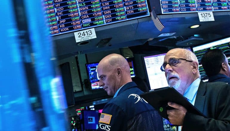 A stock trader in glasses and a suit looks up at screens showing stocks