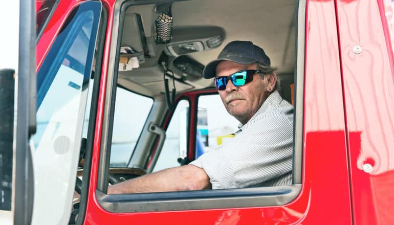 A driver with sunglasses and a ballcap on looks out the window of red truck towards the camera