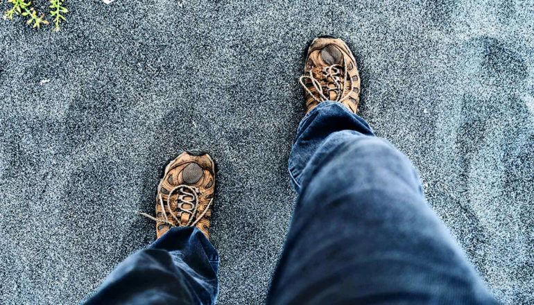 A person in gray jeans with hiking shoes on walks over gray sand