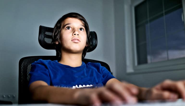 A young teen boy sits in a computer chair with his hands on a keyboard and his eyes focused