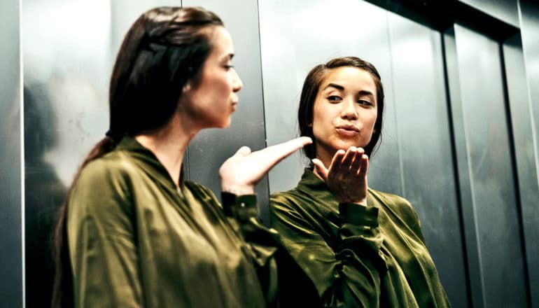 A young woman in green blows a kiss to her own reflection in an elevator mirror