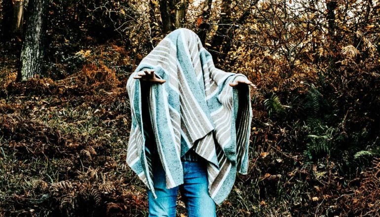 A person walks through the woods covered in a blanket with their hands outstretched