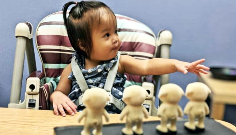 A baby girl reaches her hand out as she sits in a high chair with four toy figures in front of her