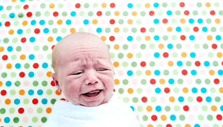 A baby cries while swaddled in a white blanket with spots of color on the blanket in the background