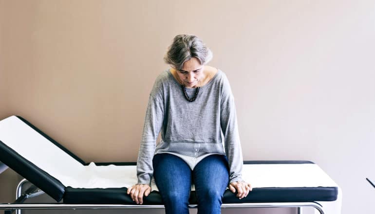 An older woman sits in the doctor's office on a table looking down