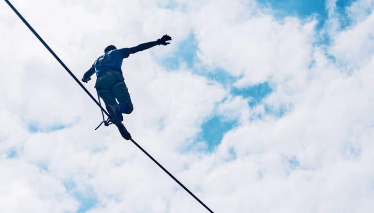 A person walks on a tightrope, balancing themselves as they go, against a cloudy sky