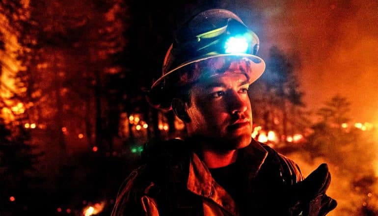 A firefighter looks off into the distance while wearing a headlamp at night during a forest fire, which casts an orange glow on the whole scene