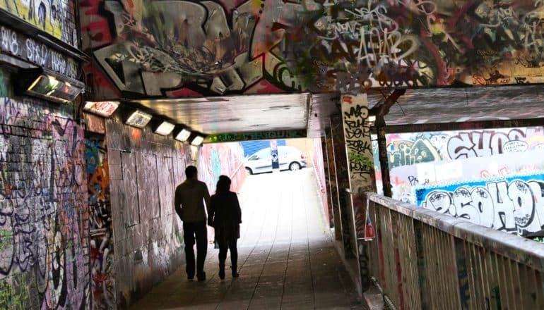 two figures walk through city tunnel with graffiti