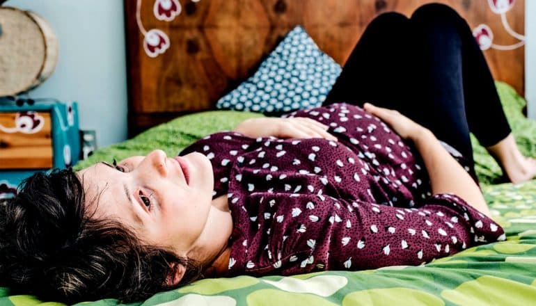 A pregnant woman lies on her bed with her head at its foot, lost in thought