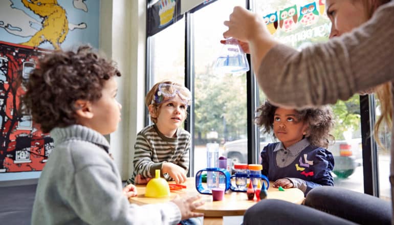 three kids in classroom look at adult holding up lab flask