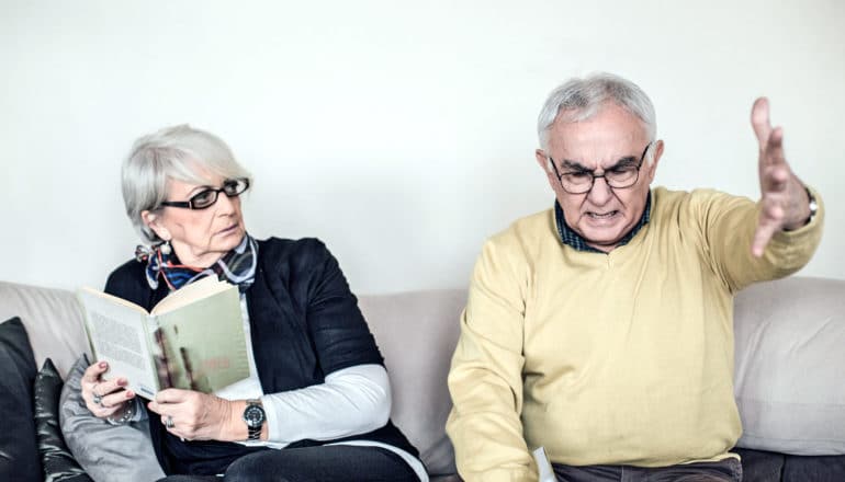 older adult holding book looks alarmed as other older adult gestures angrily