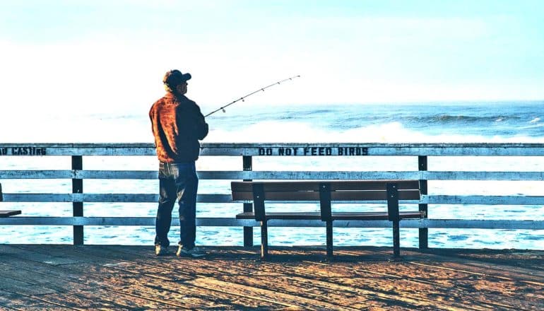 A man on a dock fishes by himself next to a bench and sign that reads "do not feed the birds"