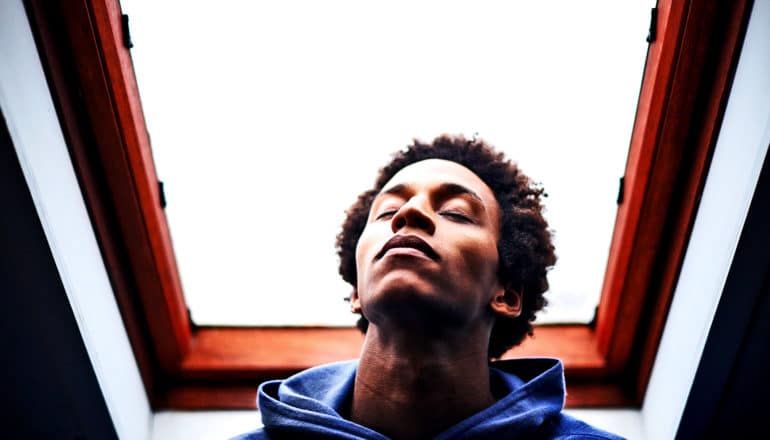 man in a blue hoodie has his eyes closed as he looks up slightly, with a skylight in the background