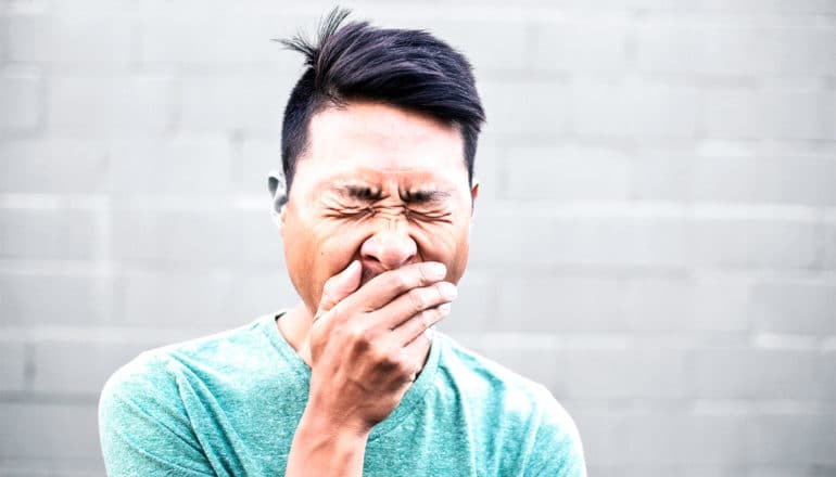 A man in a green shirt yawns deeply while standing in front of a white wall