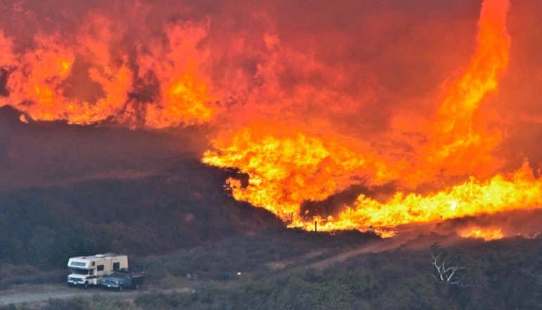 a massive wildfire burns out of control as a car and camper sit on a road in the lower left corner, showing up tiny against the giant fir