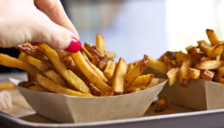 A person with a painted thumbnail reaches for a french fry from a cardboard container