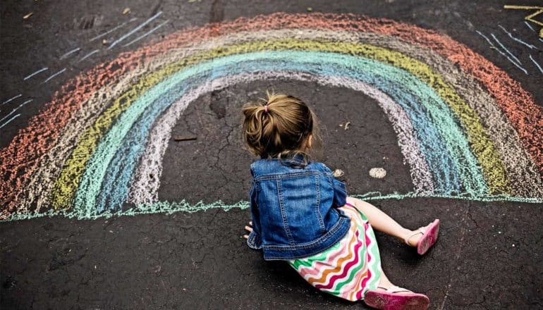 a young girl sits on asphalt drawing a rainbow with chalk