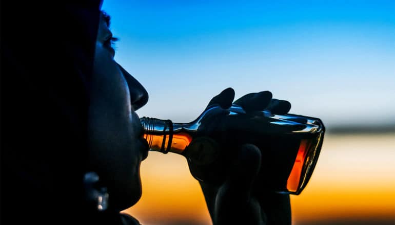 A person drinks from a small bottle of liquor as the sun sets in the background