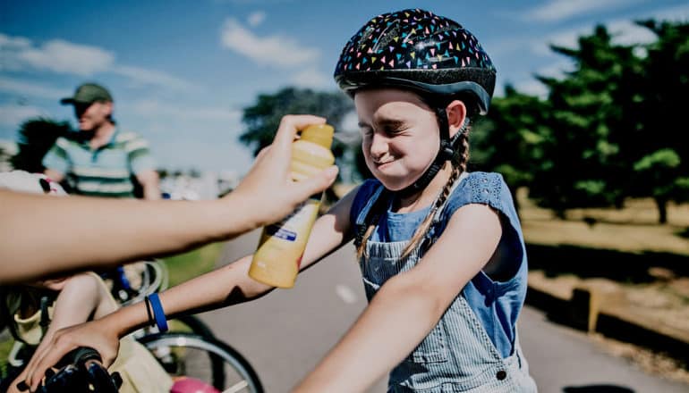 A person off-camera sprays a kid on a bike in the face with sunscreen while she grimaces